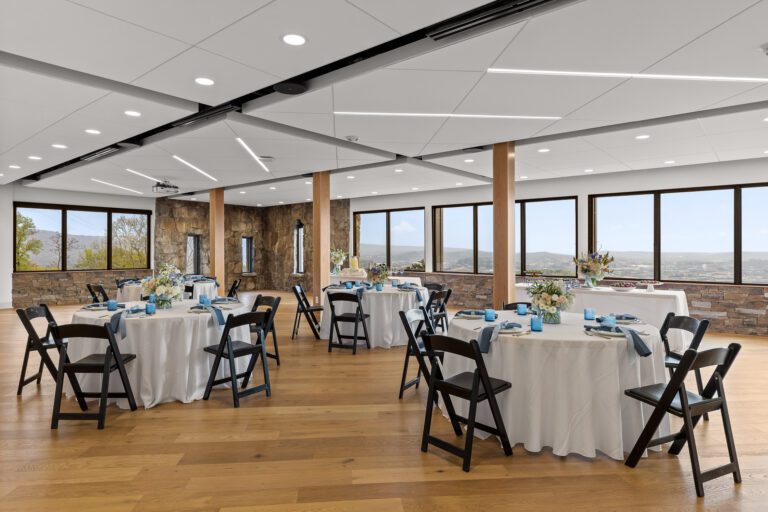 Round tables with flowers are set up in a large event room featuring walls of windows with a view of rolling hills and mountains.