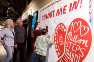 Man and woman, stand indoors in front of large banner while teenage girl and tween boy sign red and white banner showing their participation in 100 Million Steps in March.