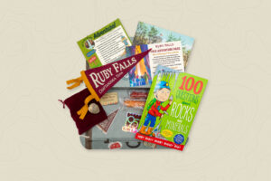 Ruby Falls Adventure Box contents with admission pass, dark red Ruby Falls vintage pennant, geode to open at home, two rock candy sticks, vintage style watercolor postcard, list of contents and blue box in the shape of a vintage suitcase.
