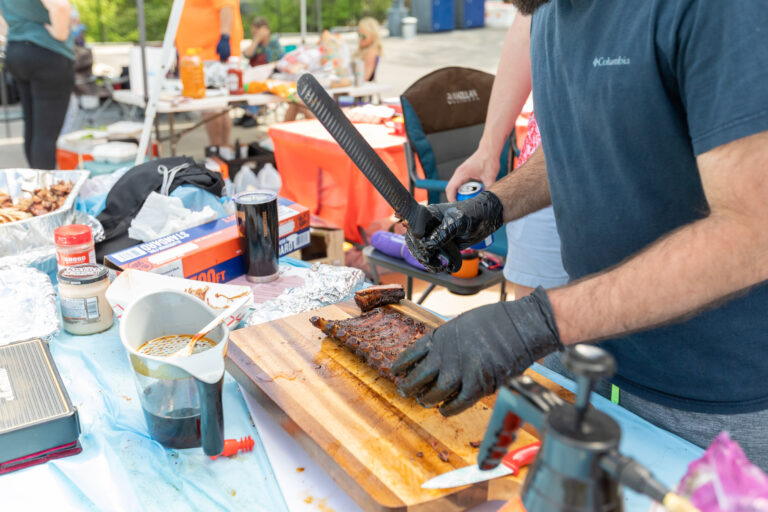 A man is cutting smoked ribs on wood cutting board outside on a table at a Battle Below the Clouds BBQ competition.