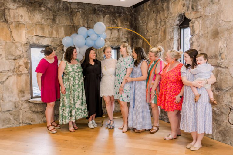 Nine women and a toddler are standing together in front of a stone wall in an event venue with a balloon arch behind them. The woman in the middle is wearing a sash that says 