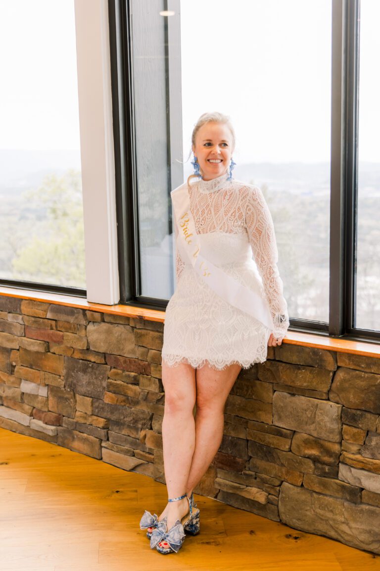 Women wearing a white lace dress stands against large windows showing a view of Chattanooga behind her.
