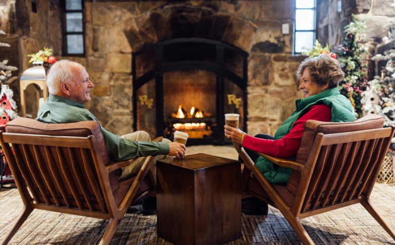 Older man and woman in seating in front of the fireplace drinking coffee.