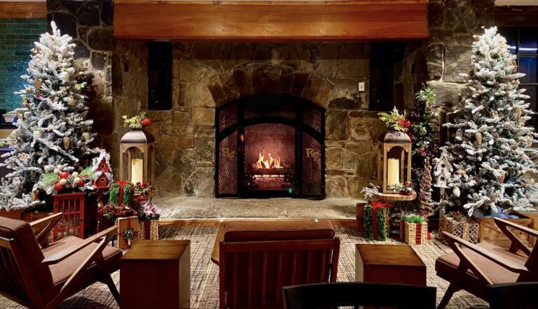 A fire burns in a stone fireplace with Christmas trees on each side featuring woodland decorations and candle lights