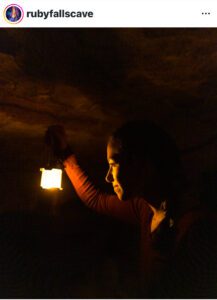 Young woman in a dark cave holding a lantern near her face.