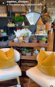 Two white chairs with yellow pillows inside a shop with shelves of plants, greenery, candles and home accessories in the background.
