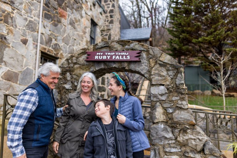 A man, woman, boy and girl stand together in front of a stone arch. A sign is attached to the top of the arch saying 