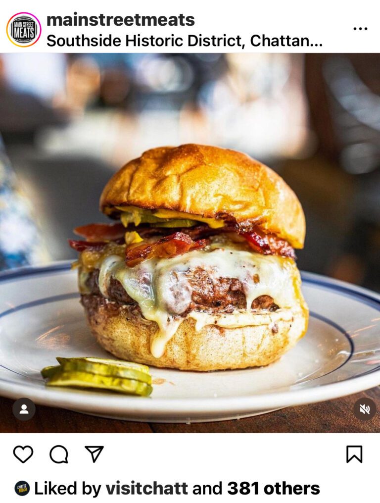 A photo shows a cheese burger with bacon topping and the top bun askew. Two pickles are on a white plate next to the burger.