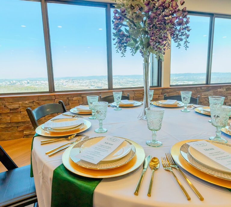 A table is set for a party with flowers, plates, napkins and glasses in Skyline Loft event venue. Large windows are in the background with a view of the Cumberland Plateau and blue sky.