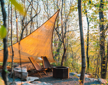 Outdoor fire pit in the woods with two chairs and an overhead shade sail.