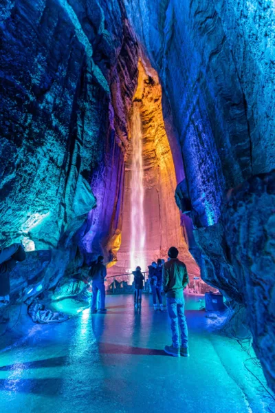 Several people stand in front of the waterfall at Ruby Falls inside the cave. Colored LED lights light up the area in blue, pink and gold. 