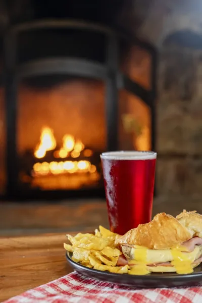 A craft beer is next to a plate with potato chips and a grilled chicken croissant sandwich is placed on a table in front of a stone fireplace with a fire. 