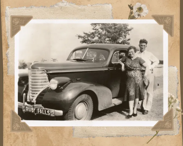 Scrapbook photo of vintage car with Ruby Falls bumper tag. A man and woman lean against the car door, smiling.