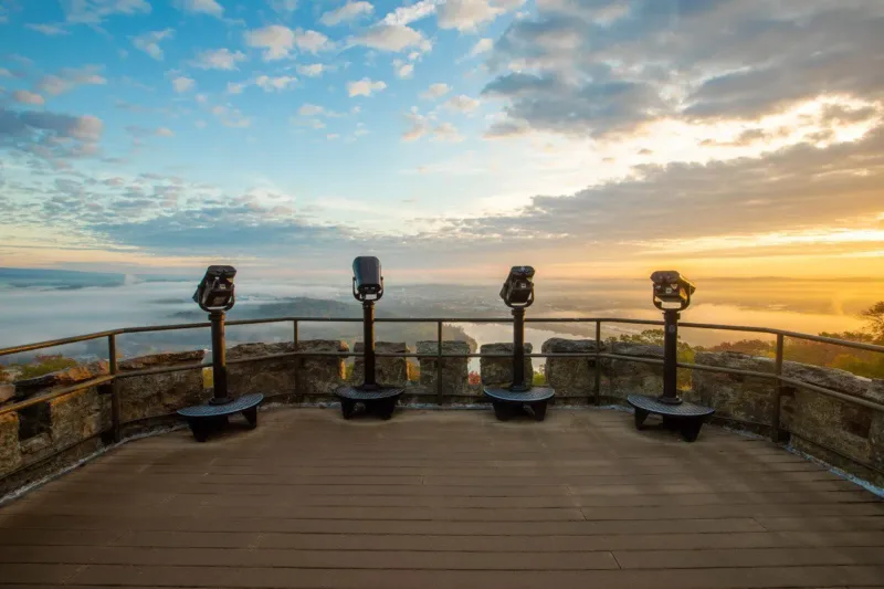 The view from historic Lookout Mountain Tower of the sun rising. The sky is filled with gold and blue hues, clouds, and golden light from the rising sun. 